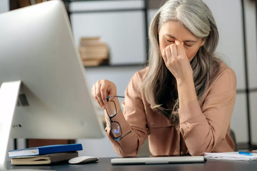Frau am PC nimmt wegen Augenschmerzen Brille ab und fährt sich durchs Gesicht